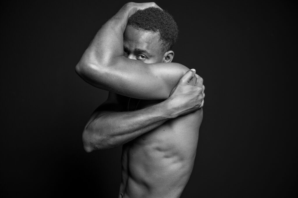 Homme torse nu en noir et blanc cachant son visage dans une posture fermée, illustrant la pression sociale et les stéréotypes toxiques liés à la masculinité.